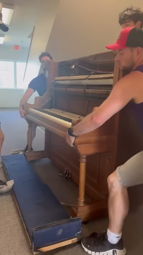 Movers in a red cap steady an open antique upright piano on a moving pad indoors