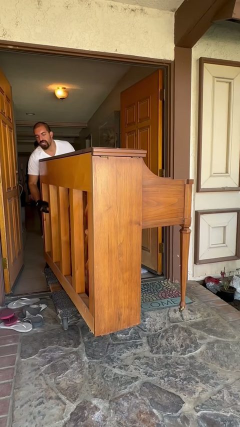 A mover carries a wooden upright piano out the front door onto a stone entry