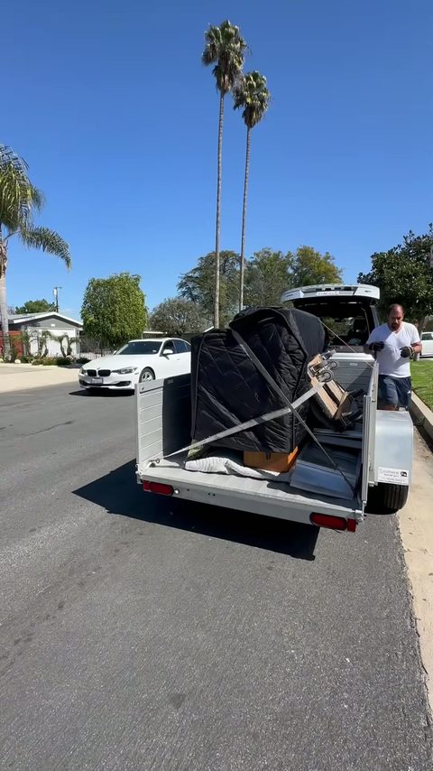 A blanket-wrapped upright piano strapped upright on a utility trailer ready for transport on a tree-lined street