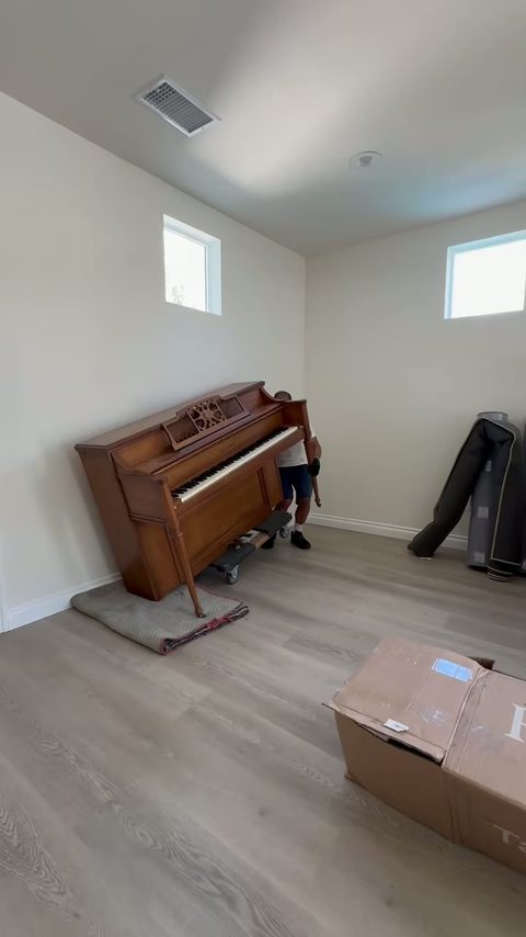 A mover rolls an antique upright piano into place in an empty new room beside a moving box