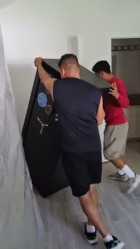 Two movers walk a tall black gun safe upright across a room