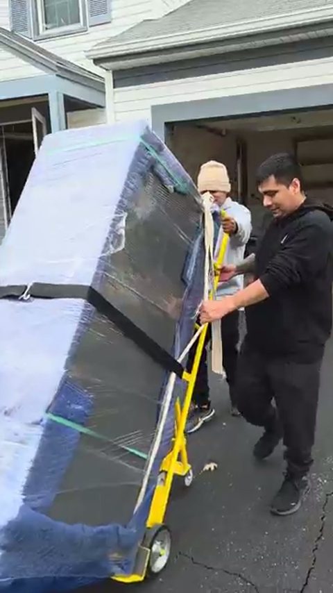 Two movers secure a blanket-wrapped upright piano on a hand truck in a suburban driveway