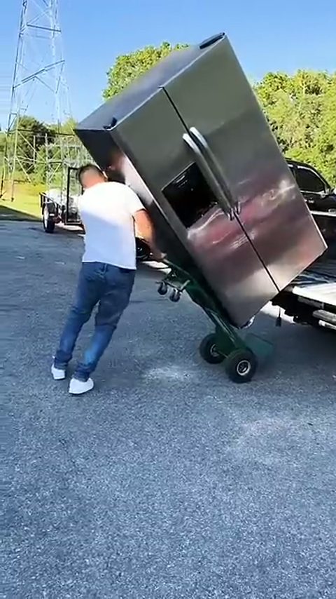 A mover tilts a large stainless steel refrigerator back onto a hand truck in an outdoor lot