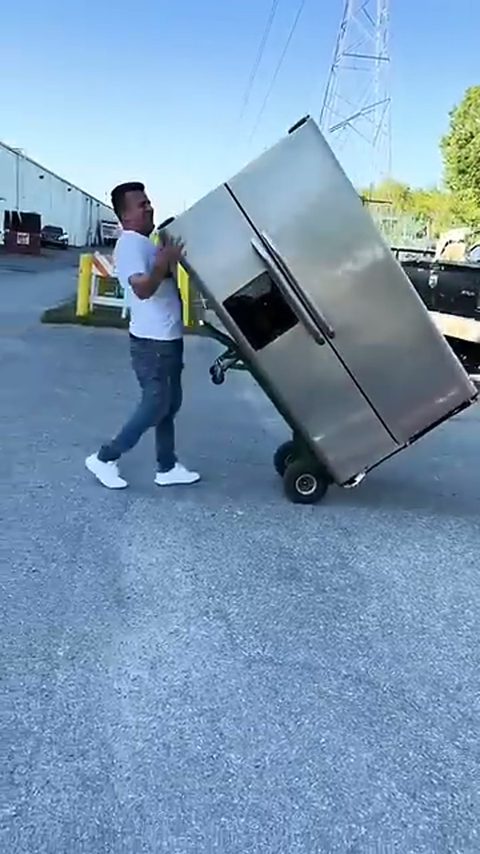 A mover wheels a stainless steel refrigerator on a hand truck across a lot
