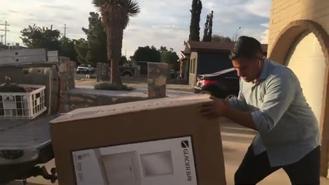 A man loads a large boxed item onto a pickup truck bed by himself outside a home