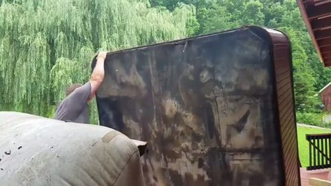 A man lifts and tilts a large hot tub on a backyard deck