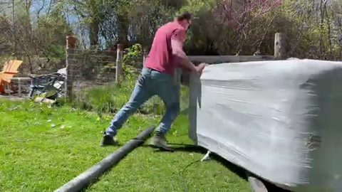 A man pushes a shrink-wrapped hot tub across a grassy yard by himself