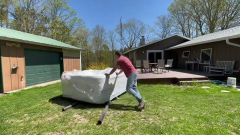 A man maneuvers a large wrapped hot tub across a lawn using pipe rollers