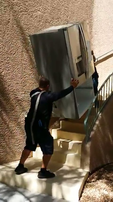Two movers carry a stainless steel refrigerator up an exterior apartment staircase